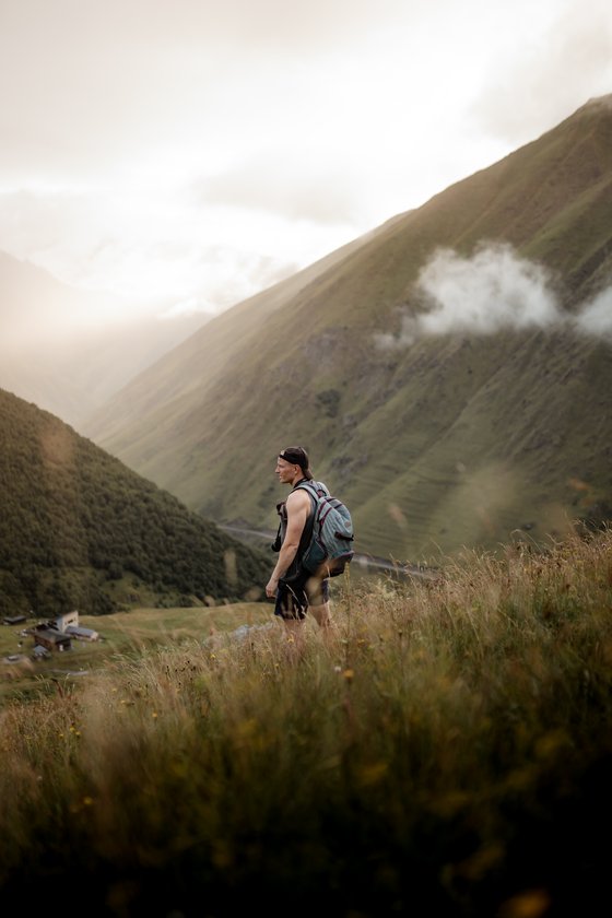 man in gray jacket and black backpack standing on green grass field near mountain during daytime