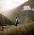 man in gray jacket and black backpack standing on green grass field near mountain during daytime