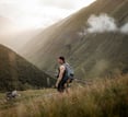 man in gray jacket and black backpack standing on green grass field near mountain during daytime