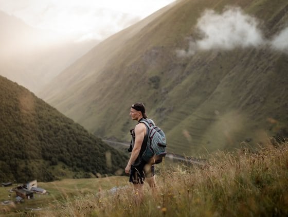 man in gray jacket and black backpack standing on green grass field near mountain during daytime