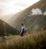 man in gray jacket and black backpack standing on green grass field near mountain during daytime