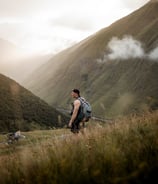 man in gray jacket and black backpack standing on green grass field near mountain during daytime