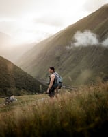 man in gray jacket and black backpack standing on green grass field near mountain during daytime