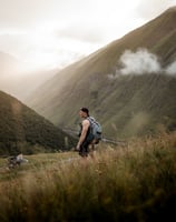 man in gray jacket and black backpack standing on green grass field near mountain during daytime