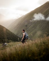 man in gray jacket and black backpack standing on green grass field near mountain during daytime