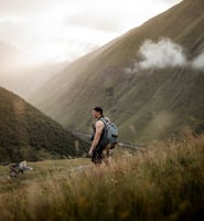 man in gray jacket and black backpack standing on green grass field near mountain during daytime