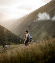 man in gray jacket and black backpack standing on green grass field near mountain during daytime