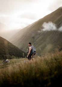 man in gray jacket and black backpack standing on green grass field near mountain during daytime