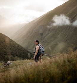man in gray jacket and black backpack standing on green grass field near mountain during daytime