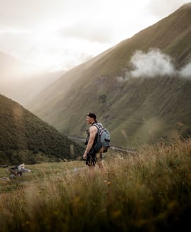 man in gray jacket and black backpack standing on green grass field near mountain during daytime