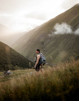 man in gray jacket and black backpack standing on green grass field near mountain during daytime