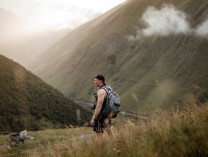man in gray jacket and black backpack standing on green grass field near mountain during daytime