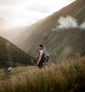 man in gray jacket and black backpack standing on green grass field near mountain during daytime