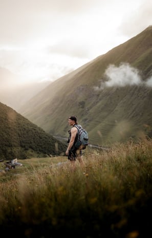 man in gray jacket and black backpack standing on green grass field near mountain during daytime