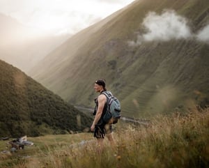 man in gray jacket and black backpack standing on green grass field near mountain during daytime