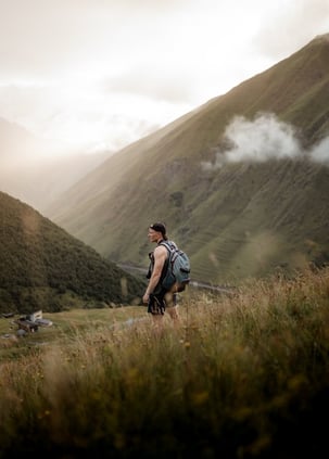 man in gray jacket and black backpack standing on green grass field near mountain during daytime