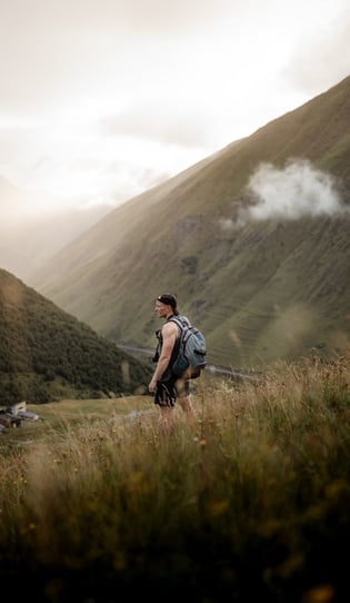 man in gray jacket and black backpack standing on green grass field near mountain during daytime