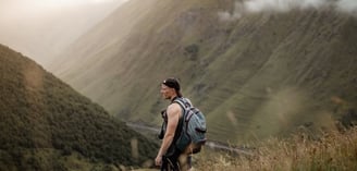 man in gray jacket and black backpack standing on green grass field near mountain during daytime
