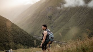 man in gray jacket and black backpack standing on green grass field near mountain during daytime