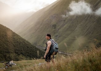 man in gray jacket and black backpack standing on green grass field near mountain during daytime