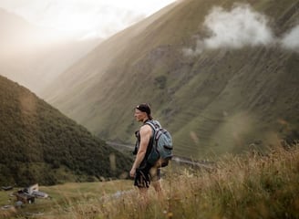 man in gray jacket and black backpack standing on green grass field near mountain during daytime