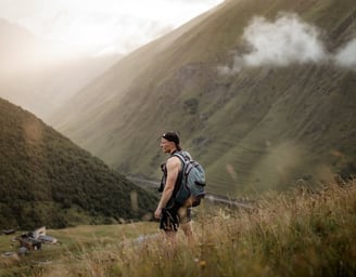 man in gray jacket and black backpack standing on green grass field near mountain during daytime