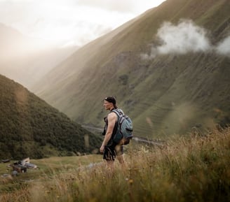 man in gray jacket and black backpack standing on green grass field near mountain during daytime
