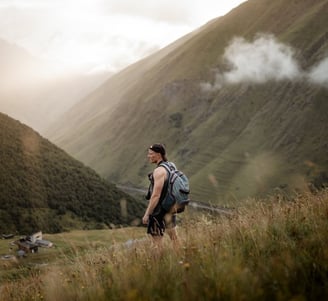 man in gray jacket and black backpack standing on green grass field near mountain during daytime