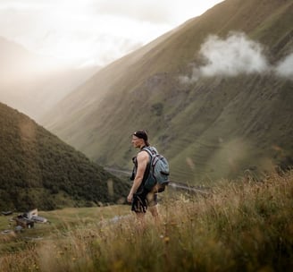 man in gray jacket and black backpack standing on green grass field near mountain during daytime