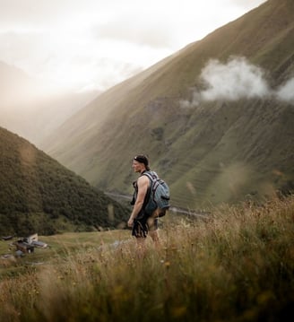 man in gray jacket and black backpack standing on green grass field near mountain during daytime
