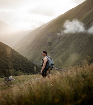 man in gray jacket and black backpack standing on green grass field near mountain during daytime