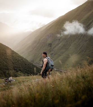 man in gray jacket and black backpack standing on green grass field near mountain during daytime