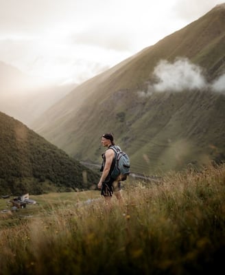 man in gray jacket and black backpack standing on green grass field near mountain during daytime