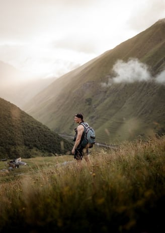 man in gray jacket and black backpack standing on green grass field near mountain during daytime