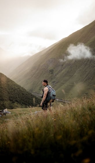 man in gray jacket and black backpack standing on green grass field near mountain during daytime