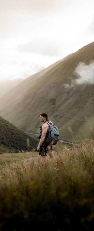 man in gray jacket and black backpack standing on green grass field near mountain during daytime