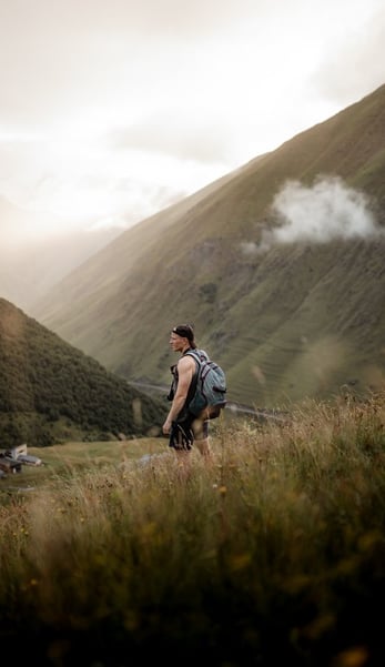 man in gray jacket and black backpack standing on green grass field near mountain during daytime