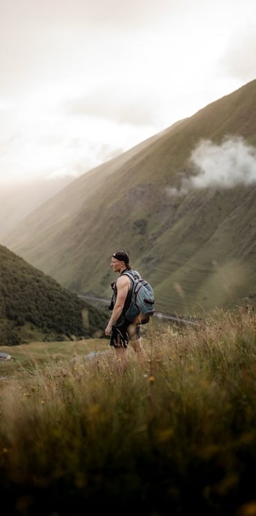 man in gray jacket and black backpack standing on green grass field near mountain during daytime