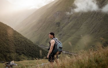 man in gray jacket and black backpack standing on green grass field near mountain during daytime