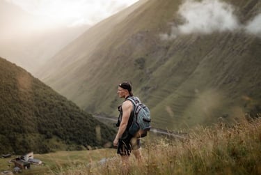 man in gray jacket and black backpack standing on green grass field near mountain during daytime