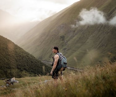 man in gray jacket and black backpack standing on green grass field near mountain during daytime