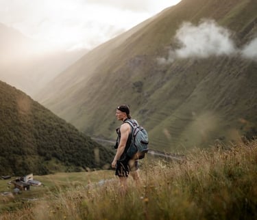 man in gray jacket and black backpack standing on green grass field near mountain during daytime