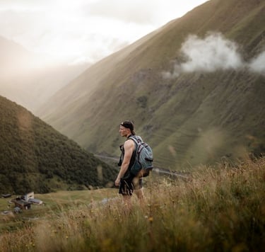 man in gray jacket and black backpack standing on green grass field near mountain during daytime
