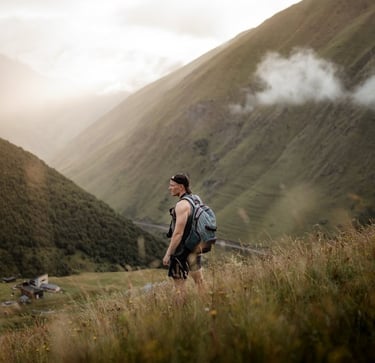 man in gray jacket and black backpack standing on green grass field near mountain during daytime