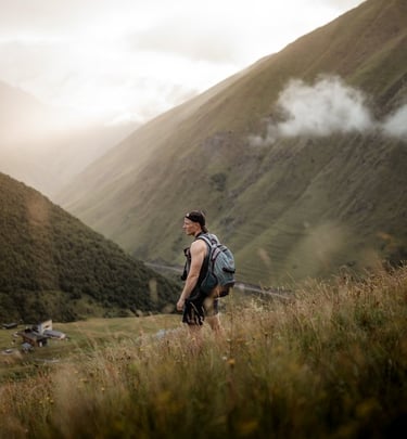 man in gray jacket and black backpack standing on green grass field near mountain during daytime