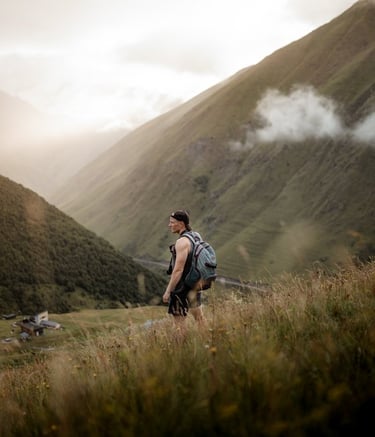 man in gray jacket and black backpack standing on green grass field near mountain during daytime