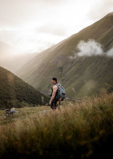 man in gray jacket and black backpack standing on green grass field near mountain during daytime