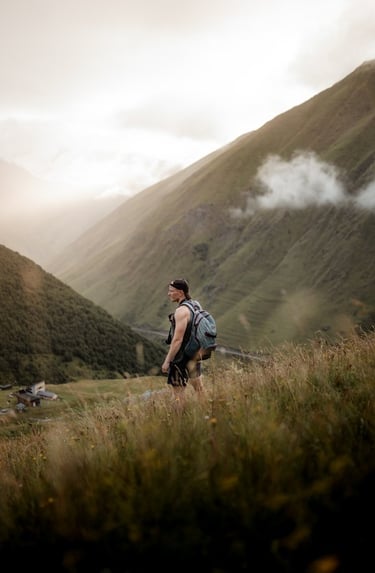 man in gray jacket and black backpack standing on green grass field near mountain during daytime
