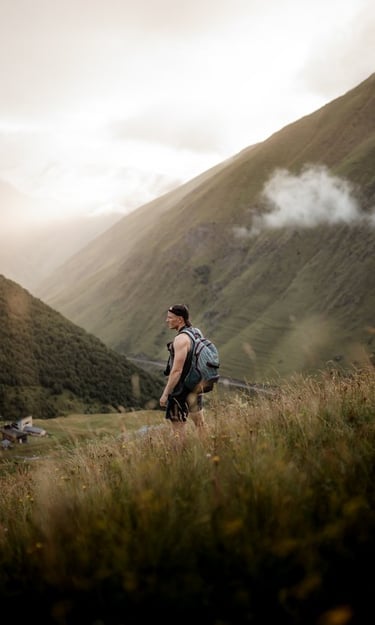 man in gray jacket and black backpack standing on green grass field near mountain during daytime