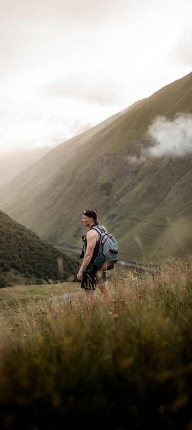 man in gray jacket and black backpack standing on green grass field near mountain during daytime